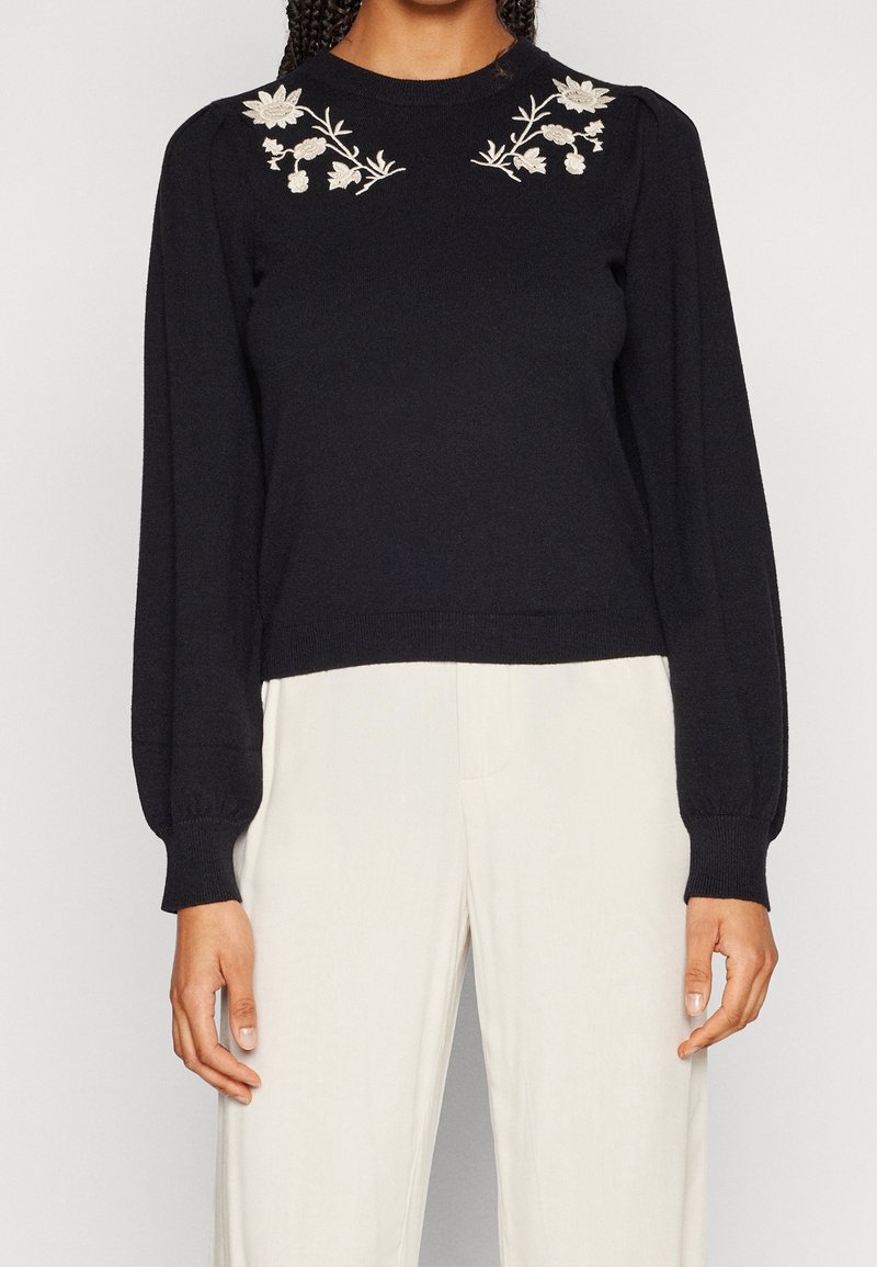 Pull en maille noire avec manches bouffantes, orné d'une broderie florale crème sur les épaules. Poignets et ourlet côtelés pour plus de texture.