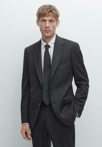 Charcoal-grauer Nadelstreifenanzug mit einem einreihigen Sakko, mit Revers und geraden Hosen. Kombiniert mit einem weißen Hemd mit Kragen und einer dunklen Krawatte.