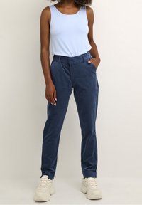 Blauwe tanktop gecombineerd met marineblauwe corduroy broek. De broek heeft zijzakken en een getailleerde pasvorm. G gedragen met lichtgekleurde sneakers.