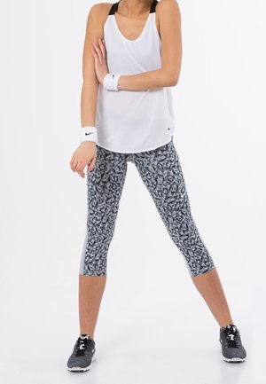 Weißes ärmelloses Tanktop mit schwarzen Akzenten, kombiniert mit gemusterten grauen Leggings und schwarzen Sportschuhen. Inklusive Armbändern.