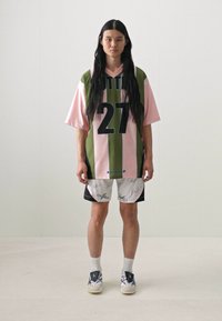 Green and pink striped oversized jersey with "27" printed. Lightweight, paired with white shorts featuring signatures. White sneakers complete the look.