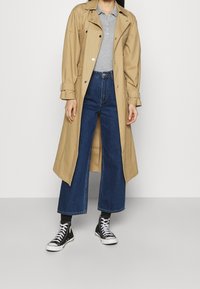 Beżowy trencz na szarym polo, zestawiony z niebieskimi dżinsami o szerokich nogawkach i czarnymi wysokimi trampkami.