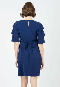 Laivansininen mekko, jossa on kerrokselliset lyhyet hihat, takana solmimisnauha ja avainaukko. Sujuva kangas ja polvipituinen leikkaus.