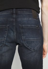 Tmavě modré džínové kalhoty s viditelným švem a zadními kapsami, které nosí osoba v černém tričku, přičemž na pravé straně je vidět část paže.