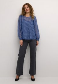 Blå, genomskinlig, texturerad långärmad topp med rund halsringning, ihop med mörkgrå, dekorerade jeans och svarta spetsiga klackskor.