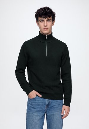 Homme portant un pull noir à demi-fermeture éclair et un jean bleu, debout avec une main dans la poche devant un fond blanc uni.