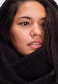 Black knitted scarf with a textured pattern wraps around the neck. Smooth skin tone and dark hair partially cover the face.