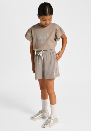 Jong meisje draagt een taupekleurig t-shirt met ruches aan de mouwen en een geometrisch ontwerp, bijpassende shorts met trekkoord, witte sokken en zilveren sneakers.