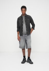 Camicia di denim nera indossata sopra una t-shirt grigia con grafica, abbinata a pantaloncini grigi. Scarpe da ginnastica nere con suola strutturata e calzini neri.