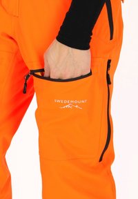 Oranje broek met een zijzak. De stof is soepel, met een ritssluiting en een wit logo met de tekst "SWEDEMOUNT" boven de zak.