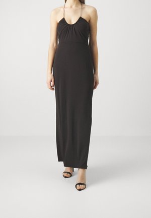 Frau trägt ein bodenlanges schwarzes Neckholderkleid und schwarze offene Sandaletten, steht vor einem schlichten hellen Hintergrund.