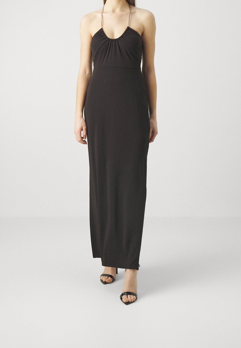 Femme portant une robe noire longue au sol avec un décolleté halter et des talons ouverts noirs, debout devant un fond clair uni.