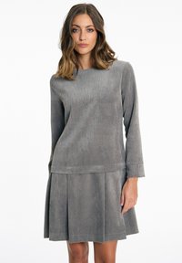 Graues Cord-Kleid mit langen Ärmeln, das eine gerippte Textur, einen runden Ausschnitt und einen weit schwingenden, gefältelten Rock aufweist. Glatte Stoffoberfläche.