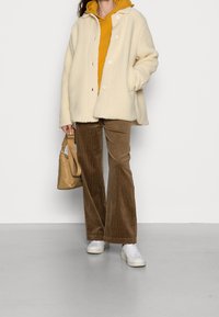 Chaqueta blanca y esponjosa sobre una sudadera con capucha amarilla, pantalones acampanados de pana marrón, zapatillas blancas, sosteniendo un bolso marrón claro. Texturas suaves y materiales blandos.