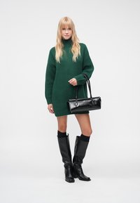 Zaļš adīts džemperkleita ar augstu apkakli, kombineēta ar ceļgalu augstiem melniem ādas zābakiem un melnu spīdīgu rokassomiņu. Vienkāršs fons.