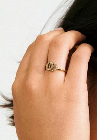 Guld ring med sammenflettede løkker prydet med små klare sten. Hånden der vises holder let om hår, hvilket fremhæver ringens design.