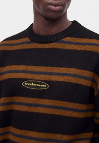 Personne portant un pull noir avec des rayures horizontales marron et bleu foncé et un logo jaune "volcom" sur la poitrine.