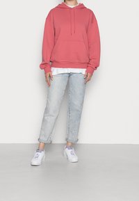 Růžová mikina s kapucí na stažení a přední kapsou, v kombinaci s světle modrými džínami a bílými teniskami. Jednoduché, neformální vrstvení s bílým tričkem.