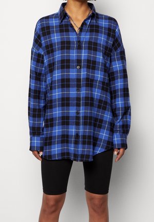 Henkilö, joka pukeutunut sinimustaruudulliseen kauluspaitaan ja mustiin istuviin shortseihin, seisoo valkoista taustaa vasten.