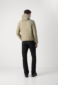 Olivnozeleni puffer jopič s horizontalnim šivi in kapuco, v kombinaciji s črnimi hlačami in črnimi čevlji, viden s hrbta.