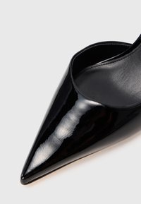 Blizgūs juodi aukšti kulnai su smailiu pirštu, su lygiavilniu odiniu vidumi ir elegantiška apdaila ant šviesaus fono.