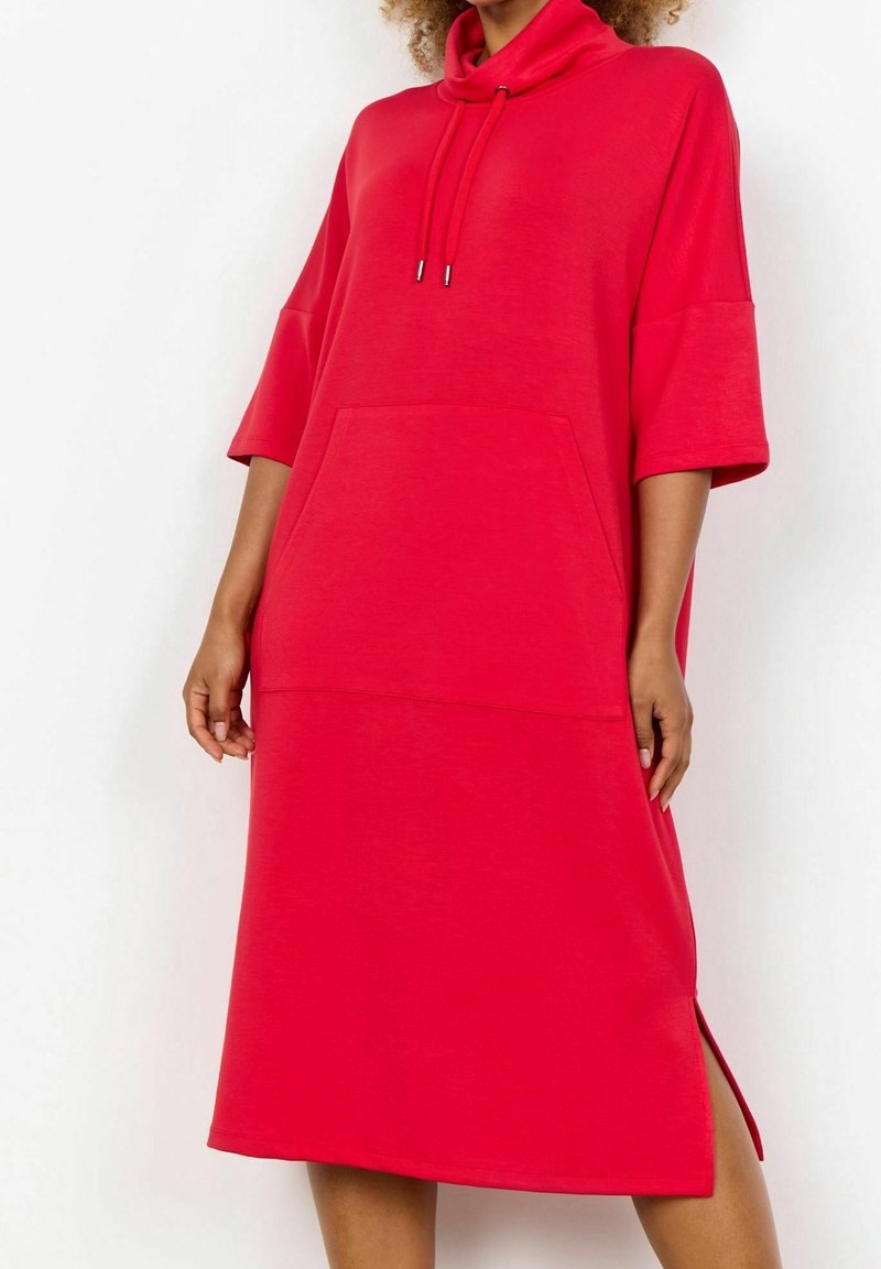 Femme portant une robe rouge ample longueur genoux avec manches trois-quarts, capuche avec cordons, poche avant, et fente latérale.