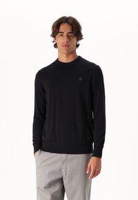 Maglione nero a maglia con collo rotondo, caratterizzato da polsini e orlo ribattuti, arricchito da un piccolo logo sul petto. Texture liscia, design aderente.