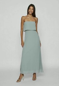 Vestido maxi verde claro, sem alças, com um corte relaxado, material fluido e comprimento até ao tornozelo, combinado com sapatos de salto dourados com tiras.