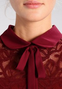 Burgunder Spitzenbluse mit einem Kragen und seidenem Bindeband. Die Spitze hat ein florales Muster, das durch eine glatte Satintextur ergänzt wird.