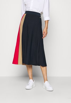 Vrouw draagt een geplooide midirok met zwarte, beige en rode panelen, een wit overhemd met opgerolde mouwen en witte sneakers.