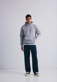 Grå hettegenser med kengurulomme, kombinert med marineblå bukser og svarte joggesko med gule såler. Jevn stofftekstur.