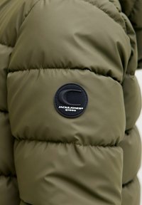 Giacca puffy verde oliva con texture trapuntata. Presenta una toppa rotonda nera con logo e testo "JACK&JONES©1990" sulla manica.