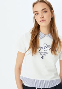 Camicia bianca a maniche corte con colletto e orlo a righe blu navy. Presenta un testo blu navy e una grafica di un'ancora. Materiale in cotone con una texture liscia.