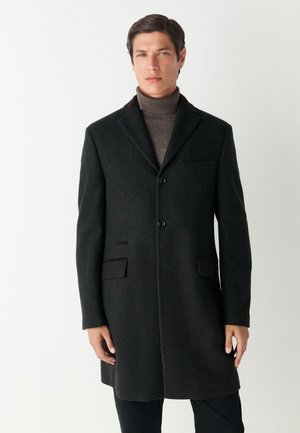 Homme portant un long manteau gris foncé charbon par-dessus un pull à col roulé marron, debout devant un fond blanc uni.