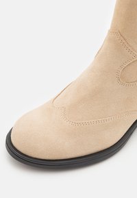 Beige sädekingad pahkluudel, sileda tekstuuriga, ümarate varvastega, musta kummist tallaga ja peene tikandiga ülaosas.