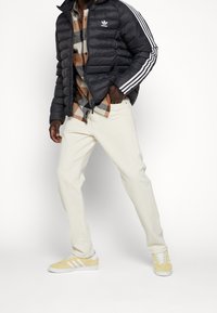 Svart puffjacka med tre vita ränder, brun och grå rutiga skjorta, krämfärgade byxor och gula Adidas-sneakers med vita accenter.