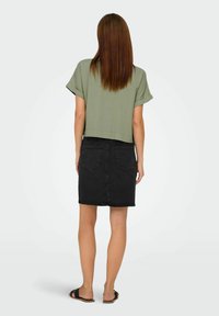 Frau mit langen braunen Haaren, die ein grünfarbenes Kurzarmoberteil, einen schwarzen Denim-Rock und schwarze Sandalen trägt, steht von hinten vor einem schlichten Hintergrund.
