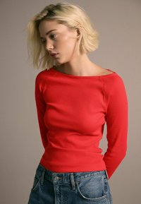 Rød, ribbet langærmet top med off-shoulder halsudskæring, parret med blå højtaljede denimjeans. Simpel, tætsiddende design.