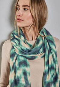 Sciarpa multicolore con un motivo ondulato nei toni del verde, blu e beige, realizzata in tessuto leggero, appesa attorno al collo di una persona.