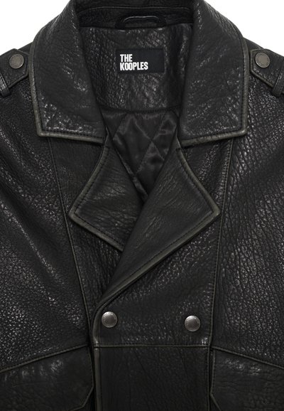 Chaqueta de cuero negro con un acabado texturizado, solapas anchas y botones de presión plateados. Presenta un forro interior acolchado. Etiqueta de la marca visible.