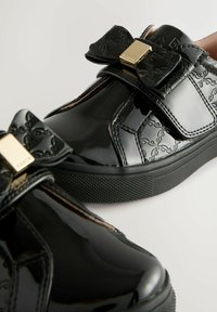 Sneakers in pelle lucida nera con un elegante dettaglio a fiocco in metallo dorato, motivo testurizzato e suola in gomma con texture.