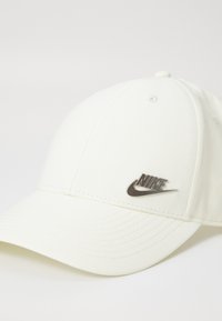 Balta beisbolo kepurė, pagaminta iš tekstūruoto audinio, su sidabriniu Nike logotipu priekyje ir išsiuvinėtais detalėmis aplink kraštą ir vainiką.