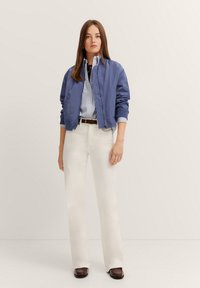 Blaue Bomberjacke mit gerippten Bündchen, über einem gestreiften Hemd getragen. Weiße Schlaghosen und braune Schuhe vervollständigen das Outfit. Neutraler Hintergrund.