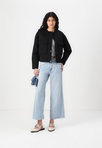 Jachetă neagră scurtă cu două buzunare frontale, purtată peste un top gri, asortată cu blugi largi de culoare bleu deschis și pantofi crem. Material texturat.