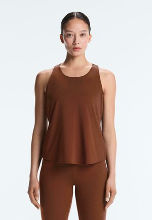 Žena so uhladeným copekom, oblečená v bezrukávovom hnedom top a zodpovedajúcich hnedých legínach na jednolom svetlom pozadí.