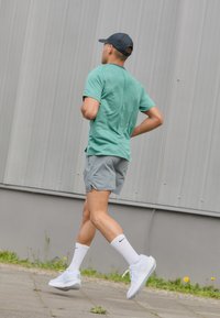 Cămașă scurtă cu mâneci scurte, de culoare verde deschis și respirabilă; pantaloni scurți gri; pantofi sport albi; șosete albe cu logo negru; persoană alergând, văzută din spate.