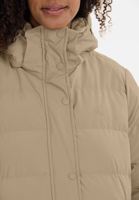 Beige puffer-takki, jossa on korkea kaulus, napit ja huomaamaton logo. Sileä kangas, jossa on tikattu rakenne.
