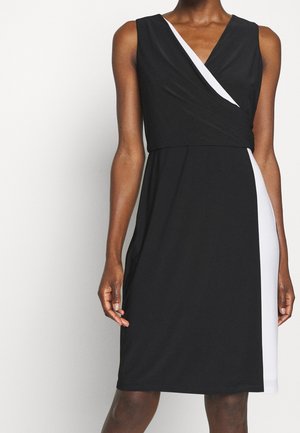 Robe noire sans manches avec un col en V, comportant un panneau blanc sur un côté et un corsage à effet portefeuille. Tissu lisse avec une coupe jusqu'au genou.