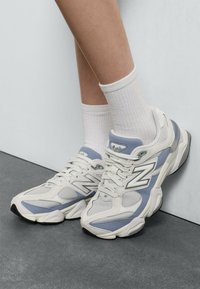 Tenisky New Balance v svetlosivej a modrej farbe, vyrobené z malých sieťových a syntetických materiálov, zaoblený tvar, polstrovaný golier a gumová podrážka.