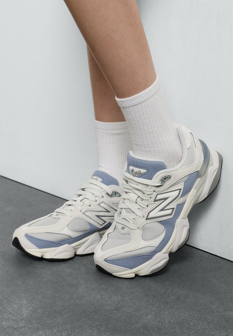 Tenisky New Balance v svetlosivej a modrej farbe, vyrobené z malých sieťových a syntetických materiálov, zaoblený tvar, polstrovaný golier a gumová podrážka.